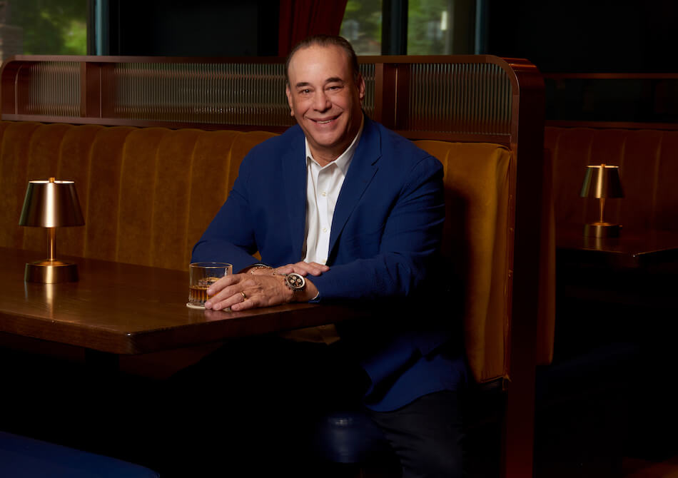 Jon Taffer in blue blazer sitting in dimly lit restaurant booth holding glass