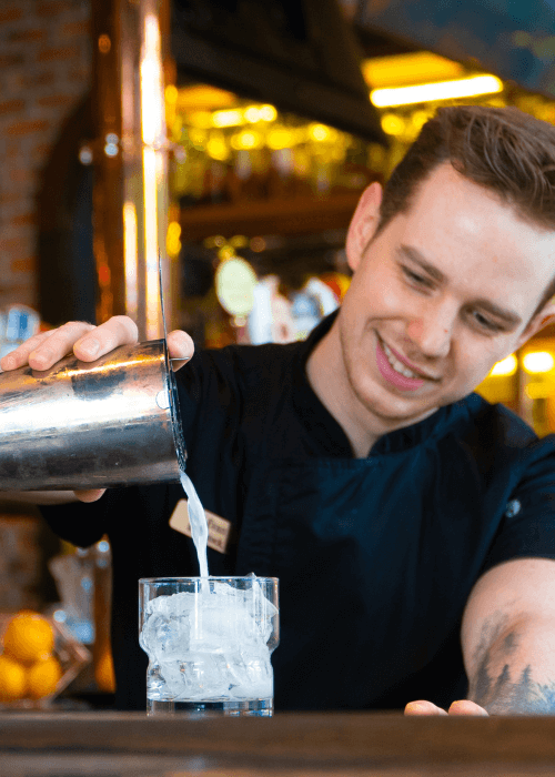 Smiling bartender pouring creamy cocktail into ice-filled glass at cozy bar
