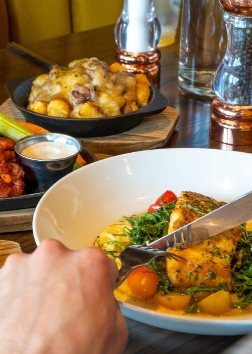 Hands holding fork and knife over grilled salmon with tomatoes and greens
